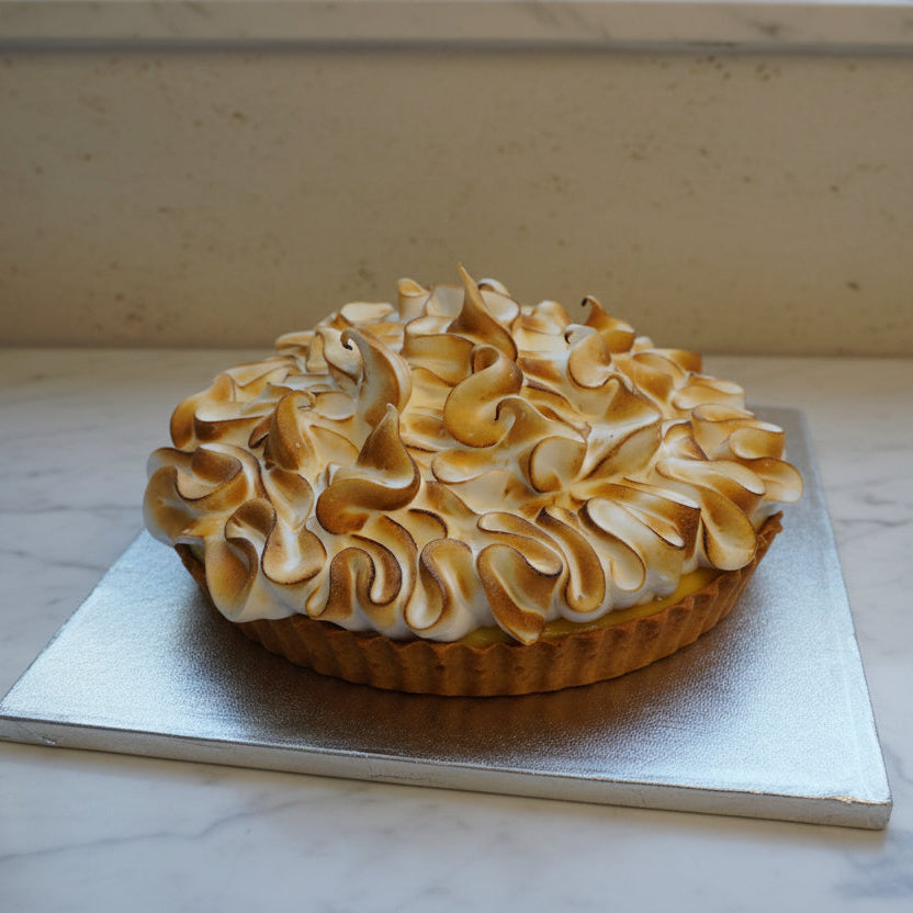 A large, beautifully 9 inches lemon meringue pie sits on a square silver cake board placed on a rustic wooden table.