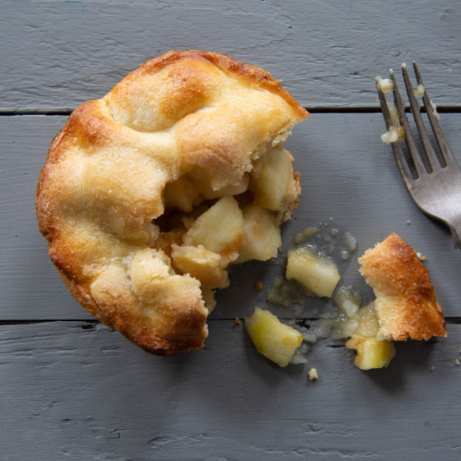 A rustic baked apple pie or crumble with a golden-brown topping and pale yellow apple pieces, shown on a dark gray wooden surface with a fork and crumbs.