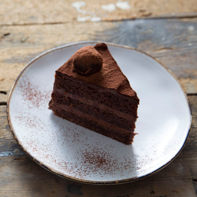 A triangular slice of chocolate fudge cake on a white plate with cocoa powder dusting, set on a rustic wooden table.