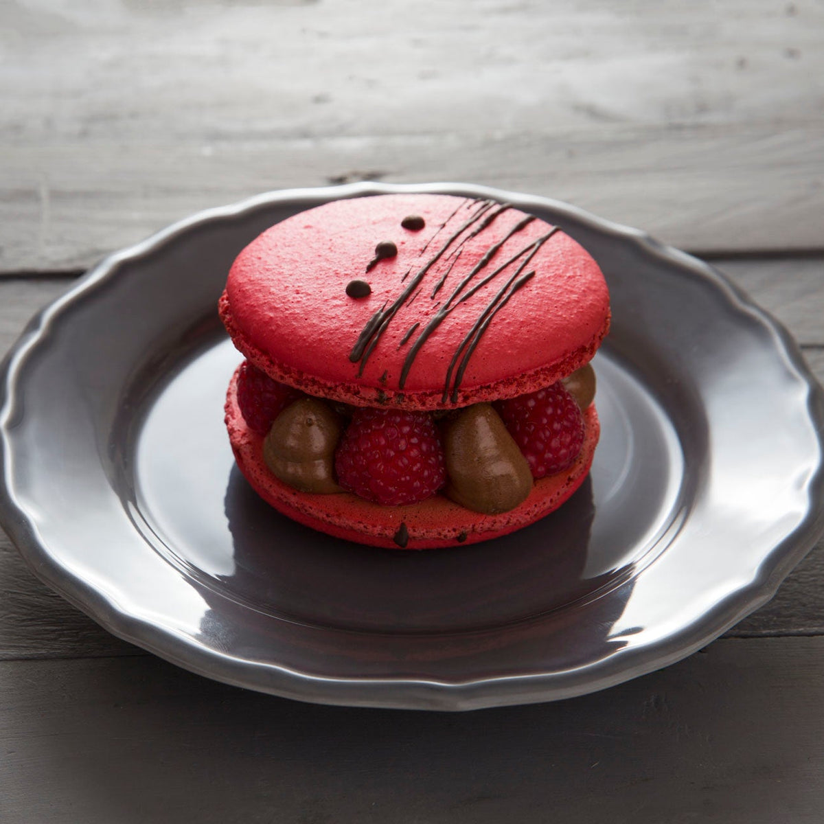 A red giant macaron with chocolate mousse and raspberry and chocolate drizzle, served on a gray ceramic plate over a rustic gray wooden table.