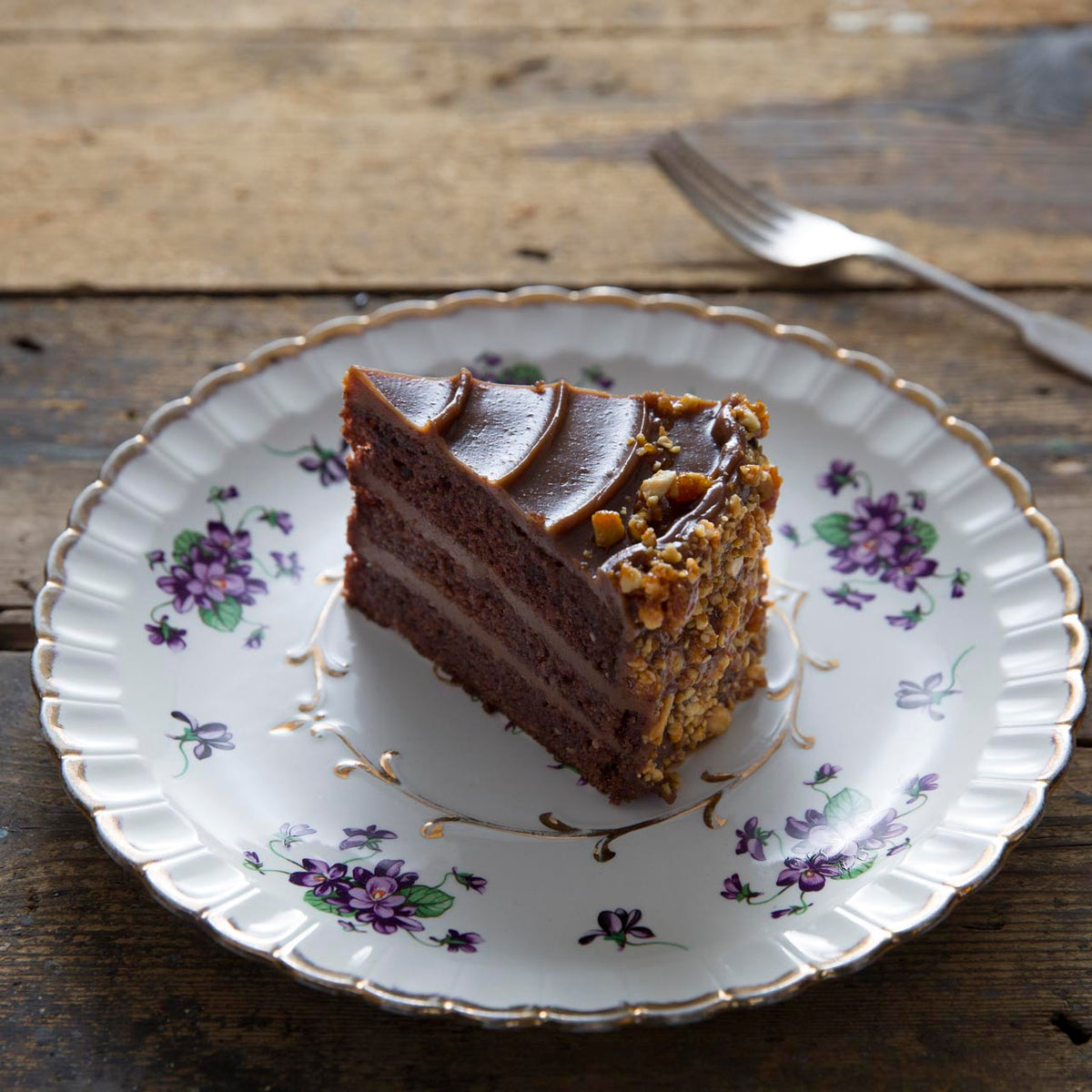 A slice of milk chocolate peanut cake sits on a white ceramic plate with purple floral patterns and gold trim, placed on a rustic wooden table with a fork in the background.