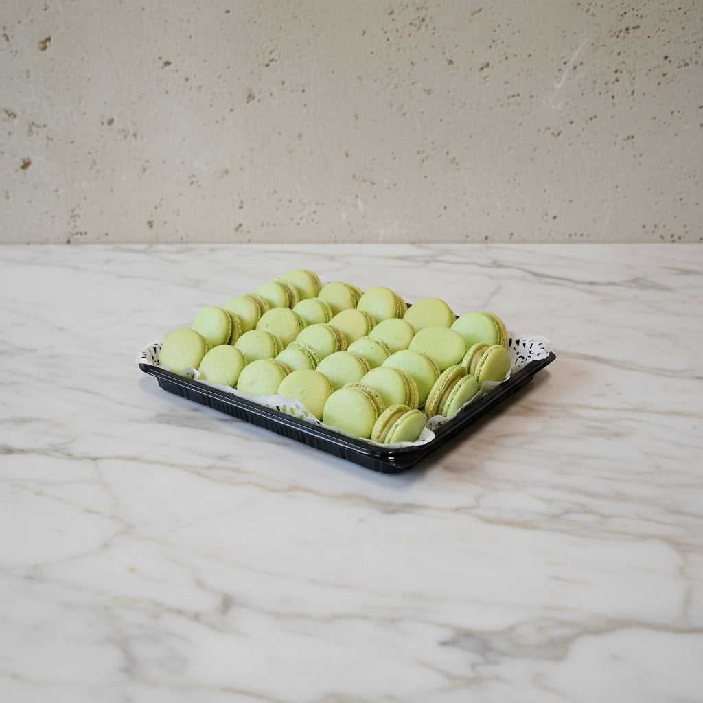 A black tray lined with white paper doilies holding rows of light green pistachio macarons, set on a stainless steel countertop.