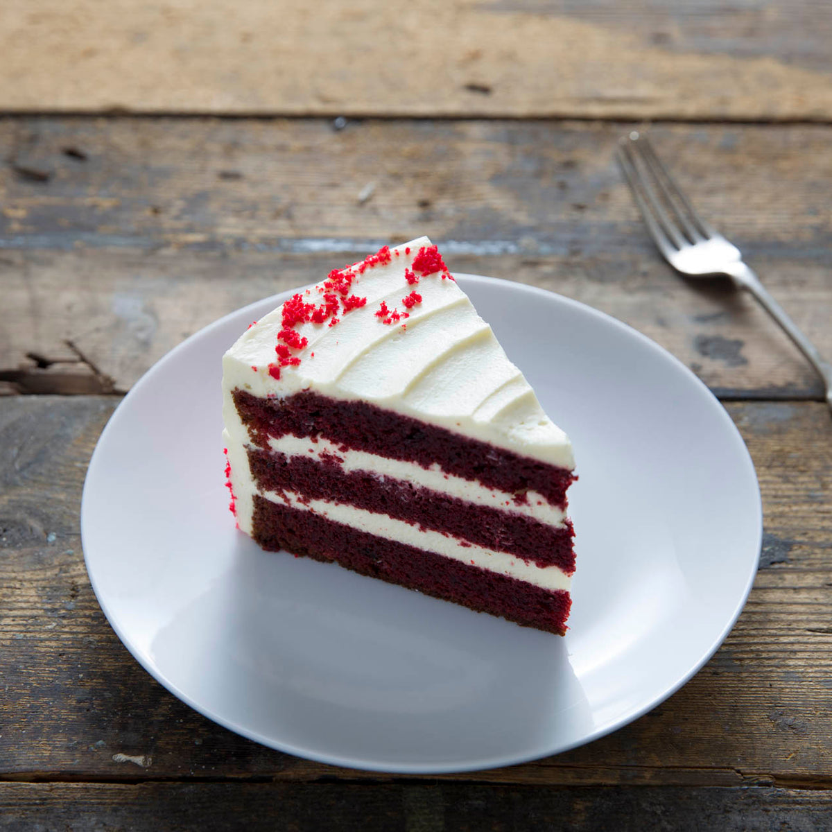 A slice of red velvet cake with white frosting on a white plate, set on a rustic wooden table next to a fork.