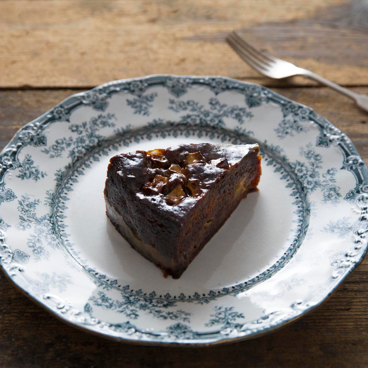 A slice of sticky toffee apple pudding, served on a white ceramic plate with a blue floral pattern, set on a rustic wooden table with a fork in the background.