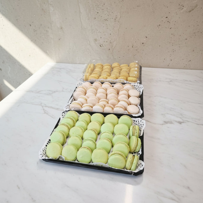 Three trays of French macarons arranged on a stainless steel counter. The top tray contains light yellow macarons, the middle tray contains pale beige macarons, and the bottom tray contains light green macarons, all displayed on black trays with white lace doilies.