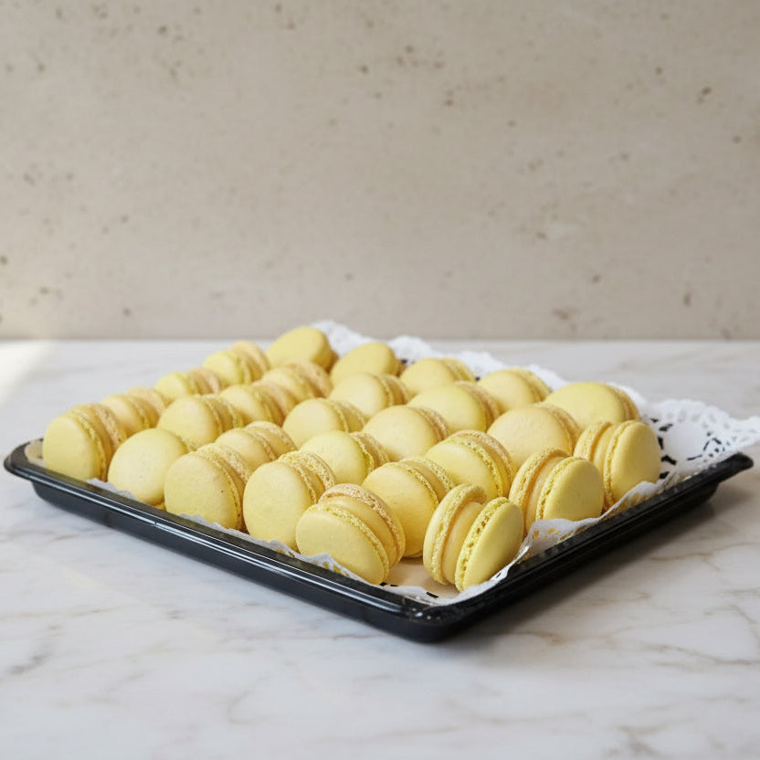 A black tray lined with a white lace doily, filled with rows of yellow passion fruit macarons, set on a stainless steel countertop.