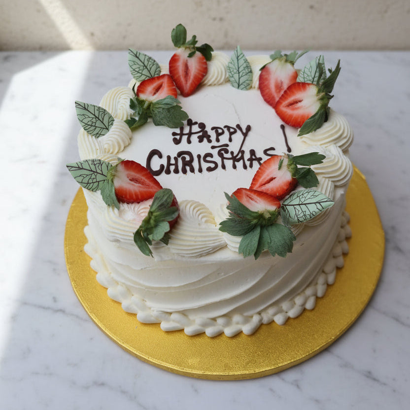 A round white frosted cake on a gold cake board, decorated with sliced strawberries and green leaves around the top, with 'HAPPY CHRISTMAS' written in icing. The cake is placed on a white marble surface against a light beige wall.