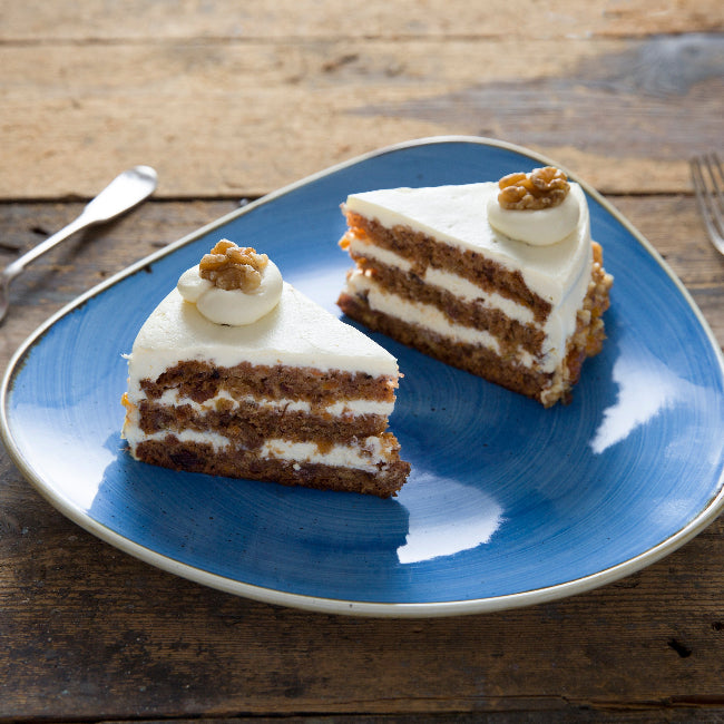Two slices of layered carrot cake with white frosting and walnut topping, served on a blue plate on a rustic wooden table with forks.
