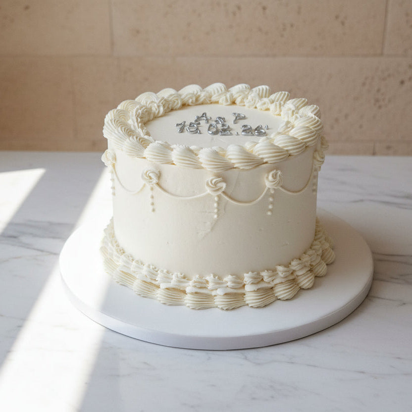 Elegant white buttercream celebration cake with piped detailing and gold lettering, displayed on a marble surface with soft natural light at Prestige Patisserie in North London.