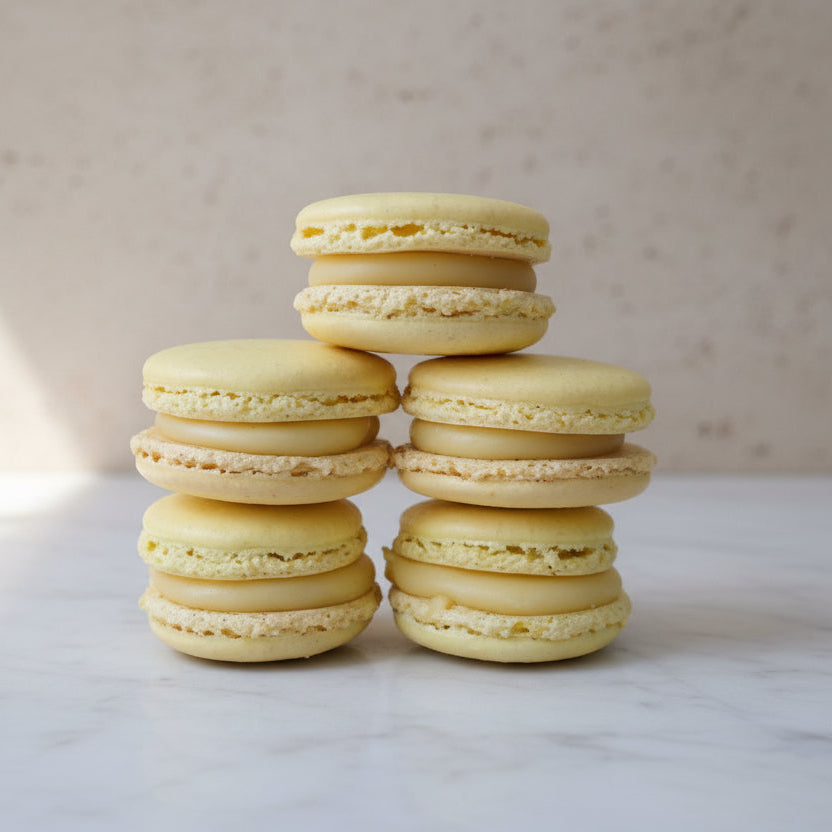 Four pale yellow macarons stacked in two columns on a rustic wooden surface, with a bamboo screen background.