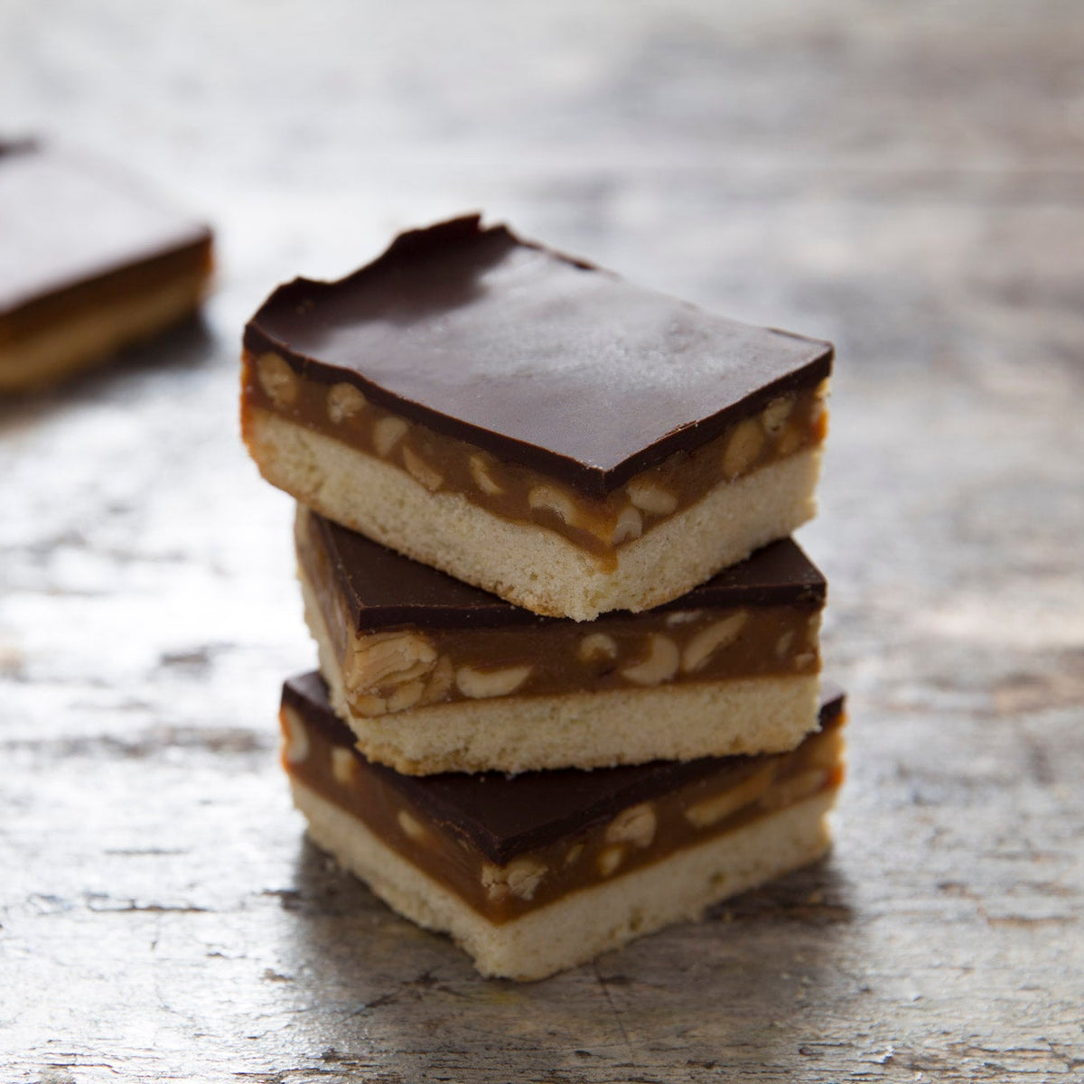 Three stacked square pieces of peanut caramel shortbread on a rustic weathered wood surface, with two more blurred in the background.