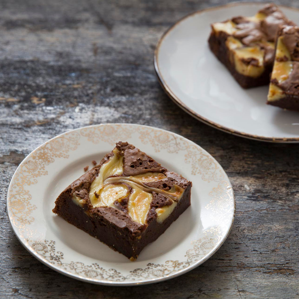 Vanilla cheesecake chocolate brownies topped served on decorative white plates with gold trim on a rustic dark wooden table.