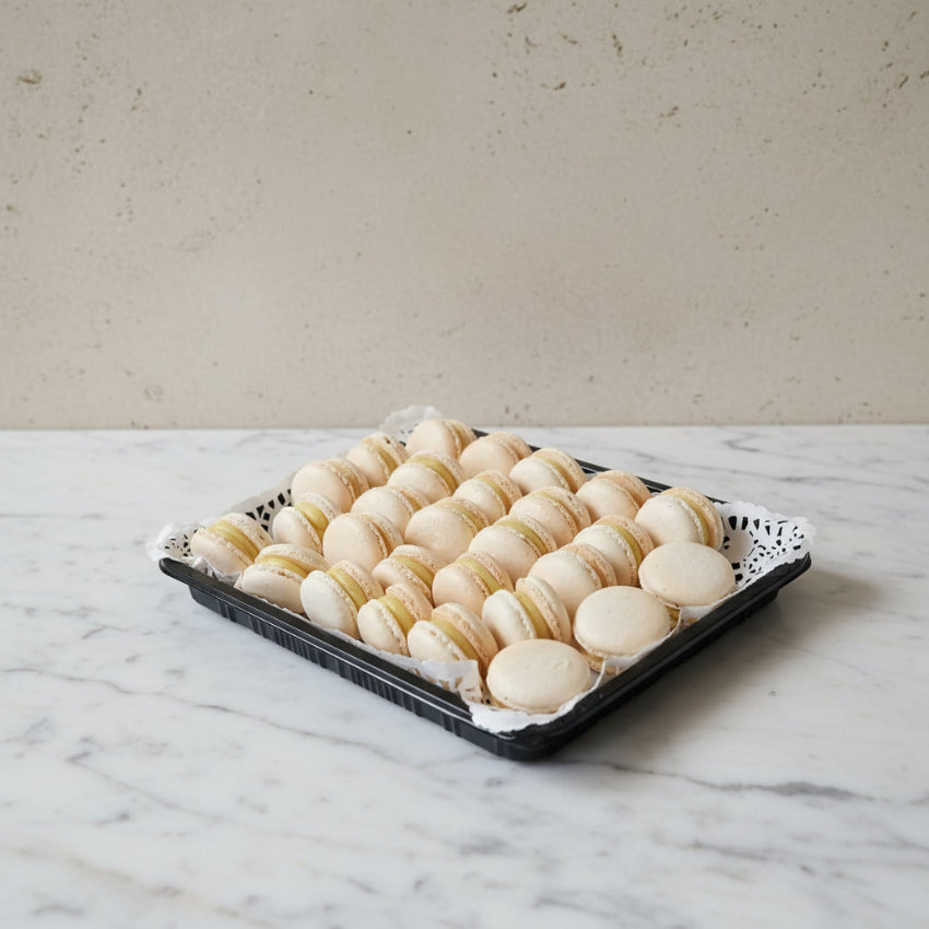A black tray lined with white paper doilies holding rows of pale beige vanilla macarons with cream filling, set on a stainless steel countertop.
