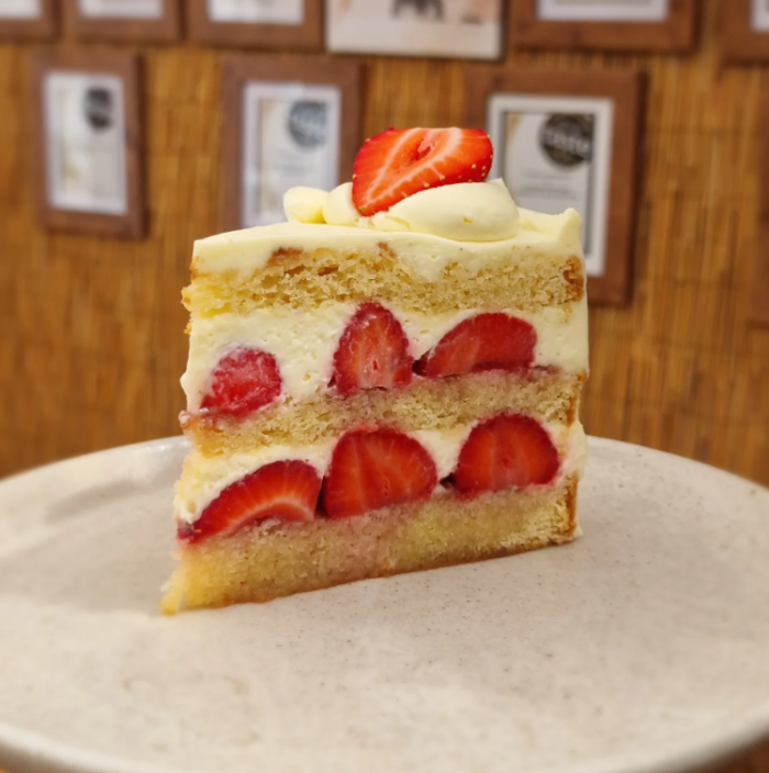 A slice of layered strawberry cake on a light beige plate, with whipped cream and fresh strawberry slices on top, set against a wooden wall with framed certificates.