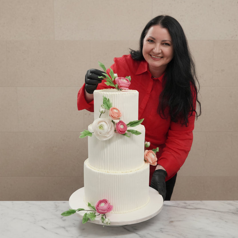 Geanina from Prestige Patisserie holding a three-tiered wedding Medovik-Honey Cake with floral decorations against a beige wall.