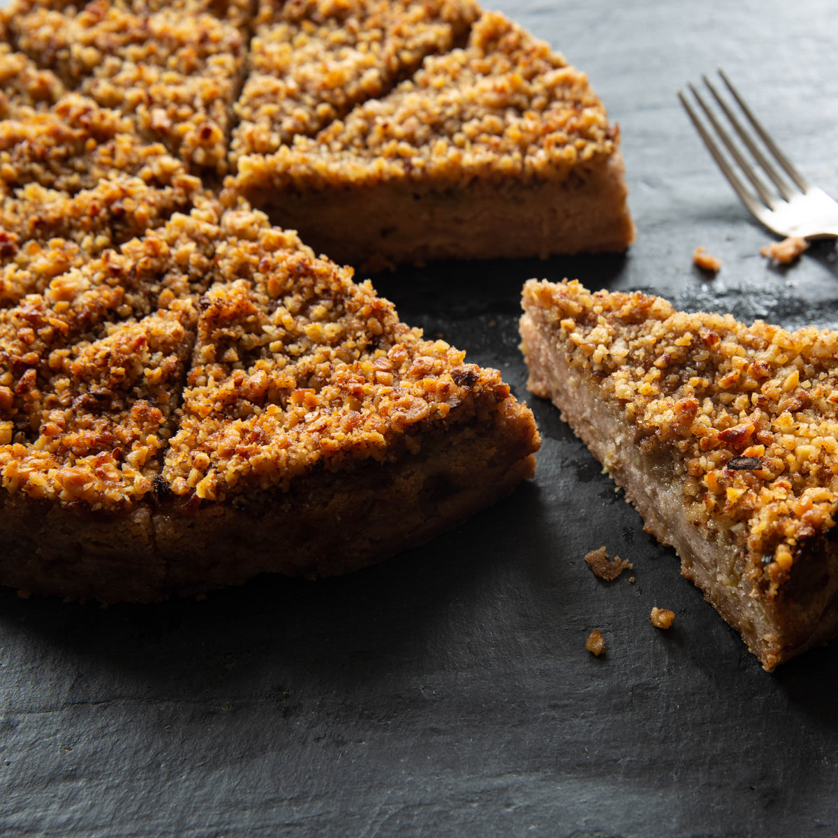 Slice of crumbly vegan and flourless apple and walnuts cake on a dark surface with a fork